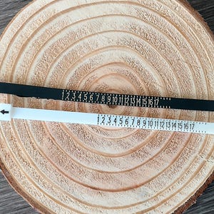 May include: Two ring sizers, one black and one white, are laid on a wooden surface. Each sizer has numbers from 1 to 17 printed on them. The white sizer has an arrow pointing to the left.