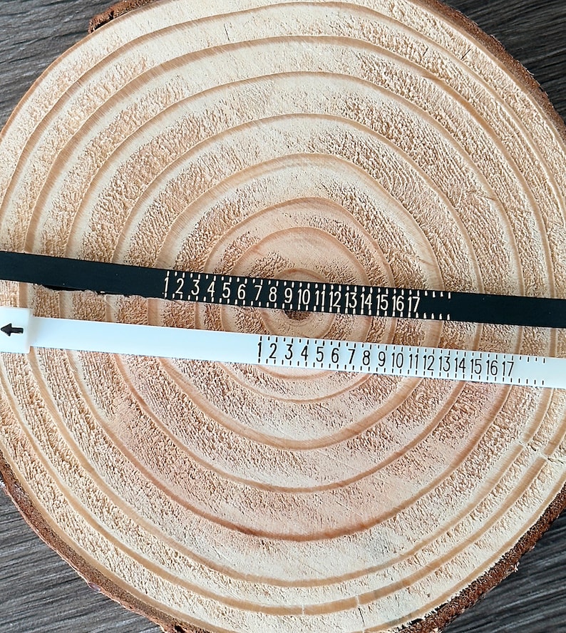 May include: Two ring sizers, one black and one white, are laid on a wooden surface. Each sizer has numbers from 1 to 17 printed on them. The white sizer has an arrow pointing to the left.