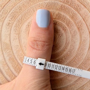 May include: A white ring sizer is wrapped around a finger with light blue nail polish. The sizer has numbers from 3 to 17, with an arrow pointing to the size. The background is a light brown, textured wooden surface.