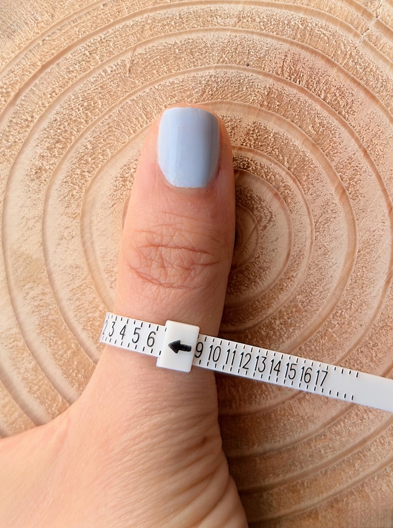 May include: A white ring sizer is wrapped around a finger with light blue nail polish. The sizer has numbers from 3 to 17, with an arrow pointing to the size. The background is a light brown, textured wooden surface.