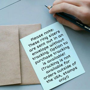 May include: A light blue sticky note with text about ring sizers and shipping information, next to a brown envelope. A hand holds a pencil in the upper right corner. The text on the note reads: "Please note: These ring sizers are sent out in an envelope without tracking."