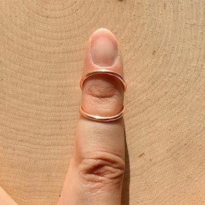 May include: A copper-colored ring worn on a finger. The ring has a simple, minimalist design, with two bands encircling the finger. The background is a light brown wooden surface.