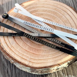 May include: A collection of ring sizers in black, white, and gray, each marked with numbers from 1 to 17. The sizers are laid out on a wooden surface, providing a clear view of the size markings for accurate ring measurements.