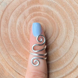 Könnte beinhalten: Ein silberfarbener Drahtring mit einem wirbelnden Design. Der Ring umschließt einen Finger und weist zwei spiralförmige Akzente auf. Der Finger ist mit hellblauem Nagellack lackiert. Der Hintergrund ist ein hellbraunes Holzmaserung.