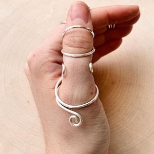 May include: A silver-colored, multi-ring finger accessory. The design features multiple bands and a decorative swirl at the base. The jewelry is worn on a hand with a light wooden background.
