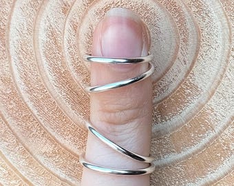 Puede incluir: Un anillo plateado se enrolla en espiral alrededor de un dedo. El anillo está hecho de un metal brillante y tiene una superficie lisa y pulida. El dedo se coloca sobre un fondo de madera con un patrón de vetas circular.
