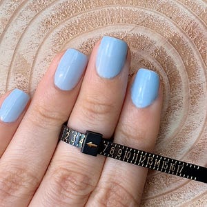 May include: A black ring sizer tool with white markings is wrapped around a finger with light blue nail polish. The tool measures ring sizes from 1 to 17. The background is a light brown, textured surface.