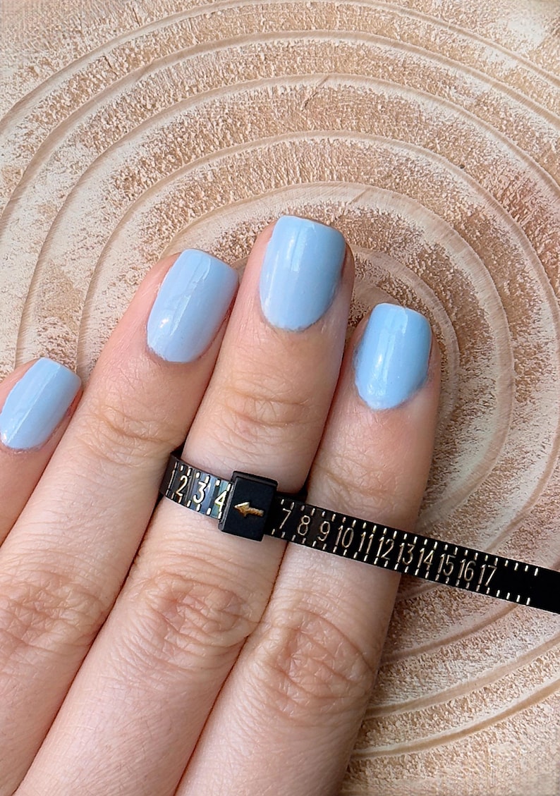 May include: A black ring sizer tool with white markings is wrapped around a finger with light blue nail polish. The tool measures ring sizes from 1 to 17. The background is a light brown, textured surface.