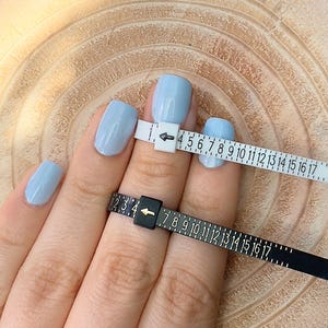 May include: A hand with light blue painted nails is shown with two ring sizers. One sizer is white with black numbers and the other is black with white numbers. The sizers measure ring sizes.