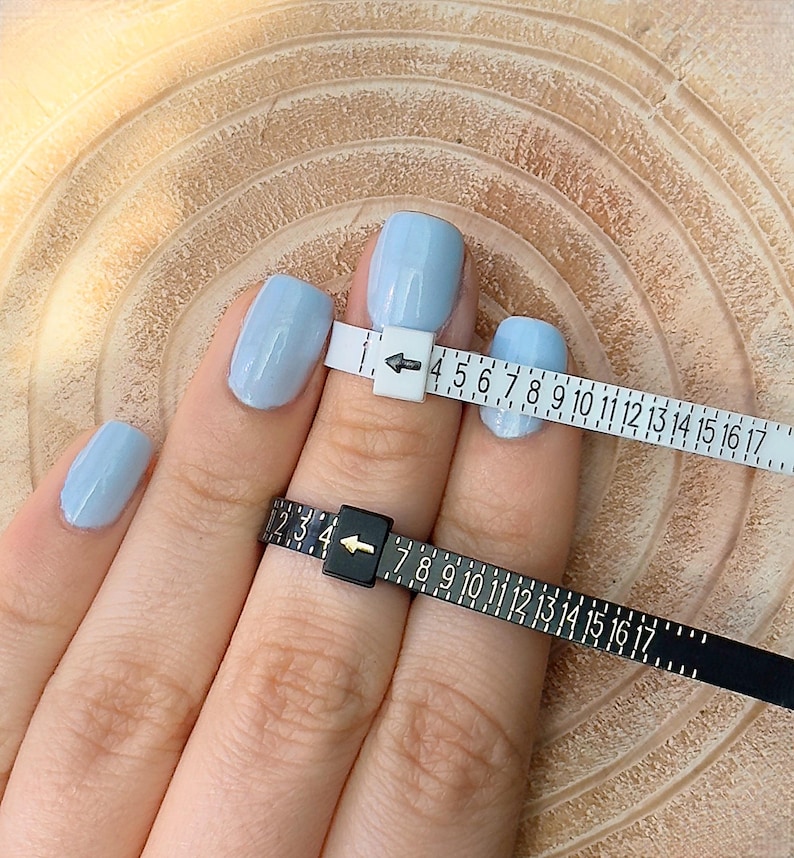 May include: A hand with light blue painted nails is shown with two ring sizers. One sizer is white with black numbers and the other is black with white numbers. The sizers measure ring sizes.