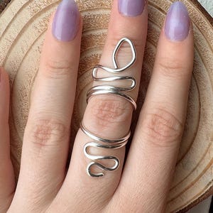 May include: A silver-colored wire ring in a snake design is worn on a finger. The ring wraps around the finger with a curved, flowing shape. The background is a wooden surface.