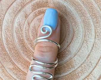 May include: A silver-colored finger ring with a unique, flowing design. The ring wraps around the finger with a series of curves and swirls, creating an elegant and artistic look. The ring is shown on a finger with light blue nail polish, set against a wooden background.