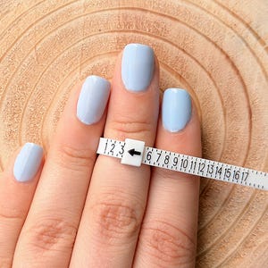 May include: A hand with light blue painted nails is shown with a white ring sizer. The ring sizer is marked with numbers from 1 to 17. The ring sizer is placed on the finger to measure ring size.