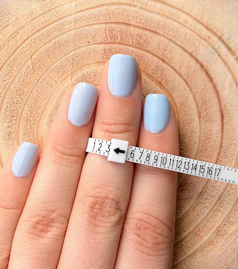 May include: A hand with light blue painted nails is shown with a white ring sizer. The ring sizer is marked with numbers from 1 to 17. The ring sizer is placed on the finger to measure ring size.