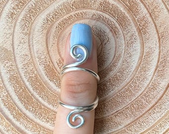 May include: A silver-colored finger ring with a spiral design. The ring wraps around the finger with a swirling detail at the top and bottom. The background is a light brown, textured surface.