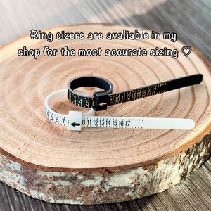 May include: Black and white ring sizers with numbered scales, resting on a wooden surface. The text "Ring sizers are available in my shop for the most accurate sizing" is visible. These tools are used to measure ring sizes accurately.