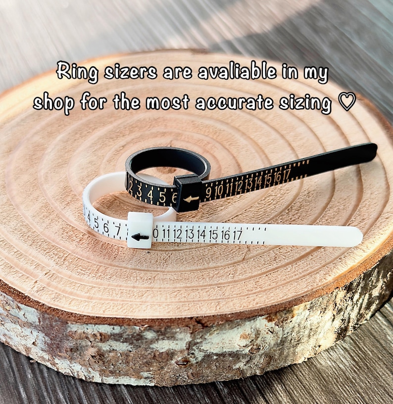 May include: Black and white ring sizers with numbered scales, resting on a wooden surface. The text "Ring sizers are available in my shop for the most accurate sizing" is visible. These tools are used to measure ring sizes accurately.