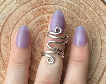 May include: A silver-colored finger ring with a swirling design is worn on a finger with a lavender-colored nail. The ring wraps around the finger with a curved, open design. The background is a light brown, wooden surface.