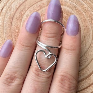 May include: A silver-colored ring with a heart-shaped design. The ring is worn on a finger with light purple nail polish. The ring's heart shape is formed by a continuous wire, creating a delicate and romantic aesthetic.