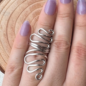May include: A silver-colored wire ring with a unique, flowing design. The ring wraps around the finger with a series of curves and loops. The ring is shown on a hand with purple nail polish. The background is a wooden surface.