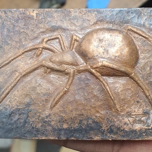 May include: A copper-colored metal sculpture of a spider with eight legs. The spider is in a raised relief on a textured background.