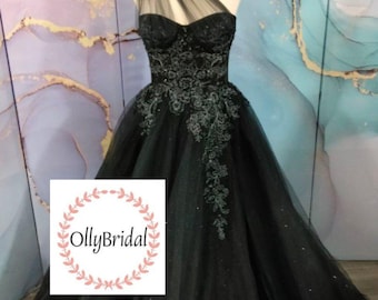May include: A black tulle ball gown with a one-shoulder neckline and silver embroidery. The dress has a fitted bodice and a full skirt. The dress is on a mannequin and the background is a light blue and purple marble pattern. The image includes the text "OllyBridal".