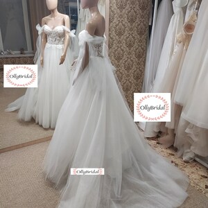 May include: A white off-the-shoulder wedding dress with a tulle skirt and lace bodice. The dress has a long train and is decorated with sequins. The dress is displayed on a mannequin in a bridal shop.