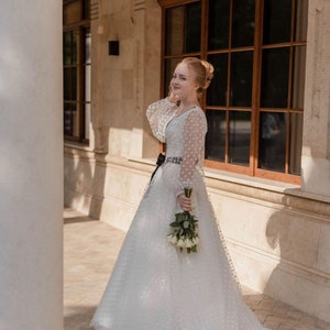 May include: A woman in a white polka dot wedding dress with a black sash and a bouquet of white roses.