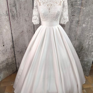 May include: A white wedding dress with a lace bodice and a full skirt. The dress has a fitted bodice with a round neckline and short sleeves. The skirt is full and flowing, with a satin finish.
