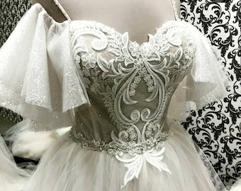 May include: A white wedding dress with a sweetheart neckline and a lace bodice. The dress has a tulle skirt and off-the-shoulder sleeves with lace detailing.