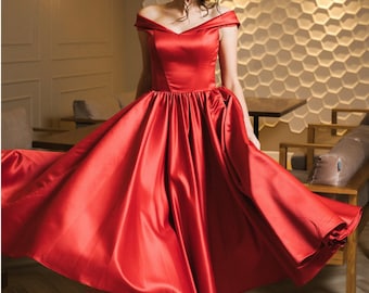 May include: A woman wearing a red off-the-shoulder satin dress with a full skirt. She is standing in a room with a white tiled wall and wooden floors.