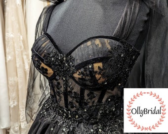 May include: A black tulle ball gown with a sweetheart neckline and a corset bodice. The bodice is adorned with black lace and rhinestones. The skirt is full and flowing. The dress is on a mannequin and has a tulle bow on one shoulder. The image includes the text "OllyBridal".