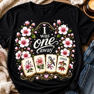 May include: Black t-shirt with the text "I was one away" surrounded by a floral design with pink and white flowers. Four playing cards are featured at the bottom, each with a different floral or bird illustration. A Joker card is also included.