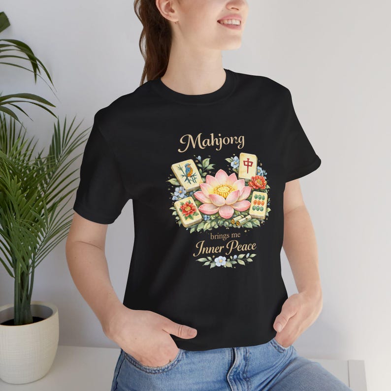 May include: Black t-shirt with a floral design and the text "Mahjong brings me Inner Peace." The design includes a pink lotus flower, Mahjong tiles, and floral elements. The shirt is made of a soft material.