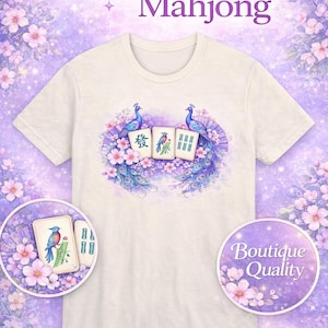 May include: White t-shirt featuring a peacock and Mahjong tile design. The design includes the text "Amethyst Mahjong" and "Boutique Quality". The shirt has a round neck and short sleeves. The background is a soft purple with floral accents.