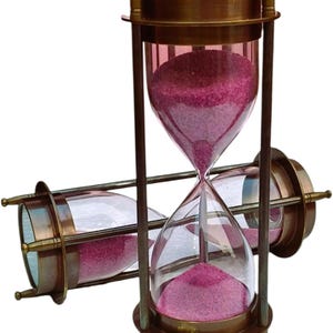 May include: A vintage hourglass with a brass frame and clear glass bulbs filled with pink sand. The hourglass is supported by a brass stand. A second hourglass is positioned horizontally behind the upright one.