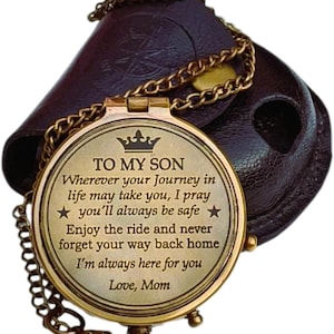 May include: A brass compass with a chain, resting on a dark brown leather case. The compass is engraved with the words "TO MY SON" and a heartfelt message from "Mom". The compass is a thoughtful gift.