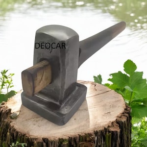 May include: A dark grey metal hammer with a wooden striking surface, resting on a tree stump. The word "DEOCAR" is printed on the hammer. The background is a blurred image of water and greenery.