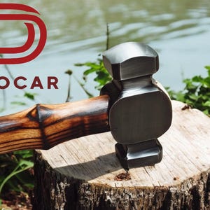 May include: A dark metal hammer with a wooden handle sits on a tree stump. The hammer's head is dark grey, and the handle is light brown with dark brown accents. The word "DEOCAR" is in red letters in the upper left corner.