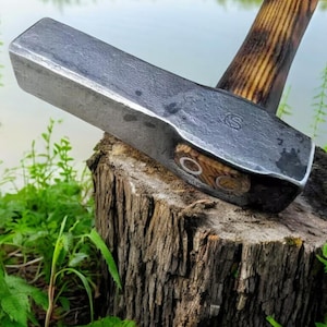 Może przedstawiać: Młot o dużej wytrzymałości z drewnianym trzonkiem. Głowa młota jest wykonana z metalu i ma fakturowaną powierzchnię. Trzonek jest wykonany z drewna i ma naturalne wykończenie.