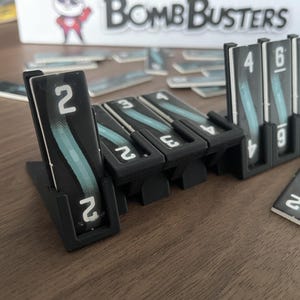 May include: Black game pieces with white numbers and a teal design are arranged on a wooden surface. The game pieces are from the board game "Bomb Busters". Some pieces are standing upright in holders, while others are lying flat.