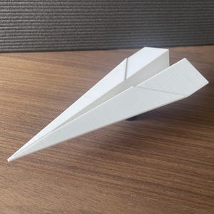 Puede incluir: Un avión de papel blanco descansa sobre una superficie de madera. El avión de juguete está hecho de papel doblado, con una nariz puntiaguda y alas anguladas. El fondo es una superficie texturizada, de color gris oscuro.