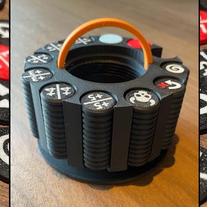 May include: A black, cylindrical game token holder with an orange handle. The holder is surrounded by black, red, and white game tokens. The tokens and holder have white symbols and numbers. The holder has a circular base.