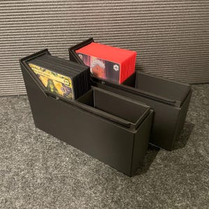 May include: Black card storage box with three compartments. One compartment holds a stack of black cards, another holds a stack of red cards, and the third is empty. The cards are visible, and the box is on a gray surface.