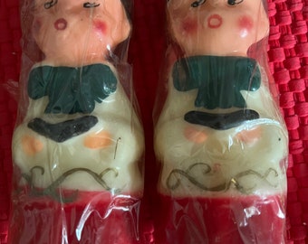 May include: Two vintage Christmas candle figurines of children wearing white robes and green scarves. The candles are wrapped in clear plastic and have red bases.