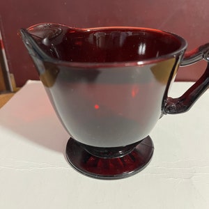 May include: A ruby red glass creamer with a decorative handle and a round base. The creamer has a spout for easy pouring. The glass is transparent, allowing light to pass through. The creamer is likely vintage.