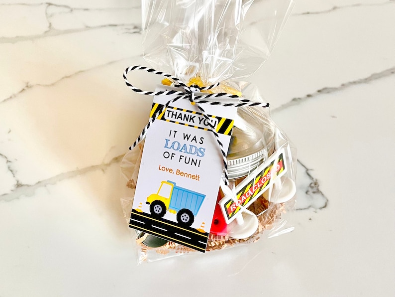 May include: A small clear plastic bag filled with construction-themed party favors. The bag is tied with a black and white twine and a tag that reads "Thank You! It was loads of fun! Love, Bennett". The tag features a yellow dump truck with blue details.