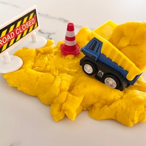 May include: A yellow play dough road with a blue toy dump truck, a red and white traffic cone, and a white sign that says "ROAD CLOSED" on it.