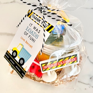 May include: A yellow and black construction themed party favor bag with a thank you tag that reads "It was loads of fun! Love, Bennett". The bag contains a small jar with a yellow lid, a road closed sign, and other construction themed toys.