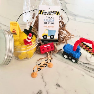 May include: A construction themed party favor with a clear jar filled with yellow play dough, a small toy dump truck, a toy excavator, a toy traffic cone, a toy shovel, a toy road closed sign, and a toy construction barrier. The jar has a silver lid. The favor also includes a thank you tag that reads "Thank You It Was Loads of Fun! Love, Bennett".